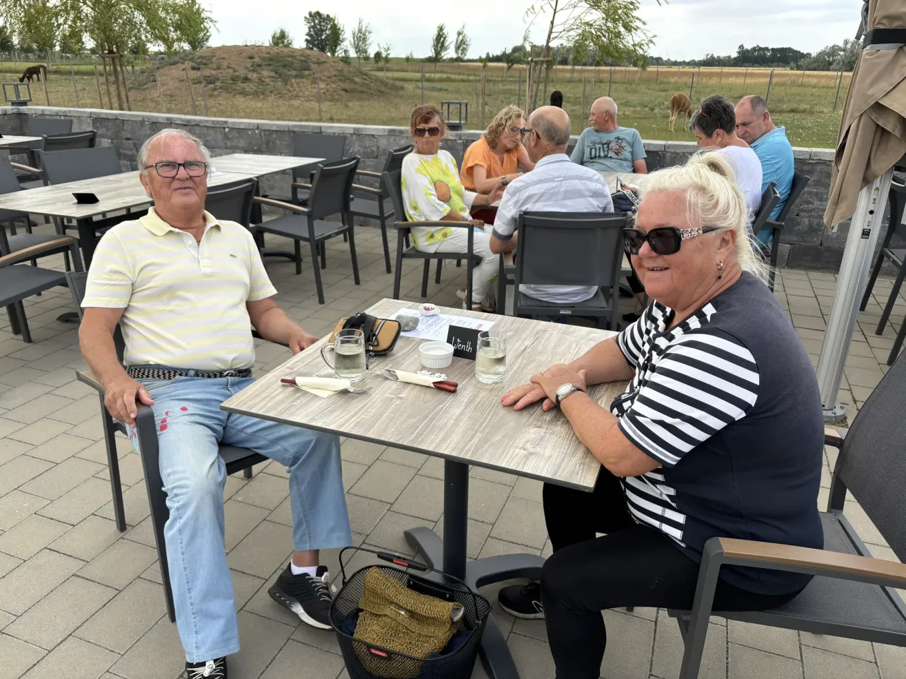 Eine Gruppe älterer Menschen sitzt um Tische in einem Outdoor-Essbereich. Ein Tisch hat eine Tafel und Gläser mit Wasser. Dahinter ist ein Grasfeld.