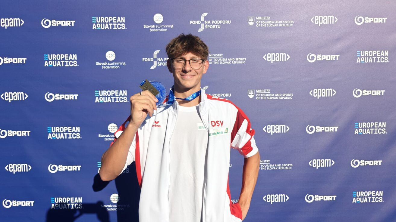 Ein junger Mann posiert mit einer Medaille vor einem blauen Hintergrund mit Logos. Er trägt eine weiße und rote Sportjacke mit den Buchstaben DSV.