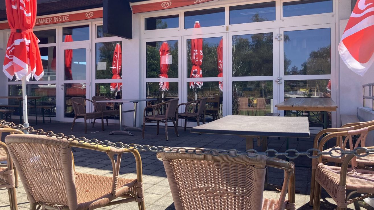 Eine Außengastronomie mit mehreren Tischen und Stühlen. Die Stühle sind aus Rattan und die Tische haben Metallbeine. Rote Regenschirme stehen auf den Tischen. Der Eingang zum Restaurant ist glasverkleidet und reflektiert die Umgebung.
