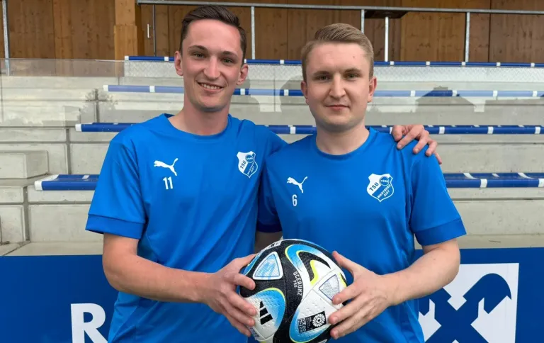 Zwei Männer in blauen Trikots stehen auf einem Sportfeld, einer hält einen Fußball, beide lächeln. Der Mann auf der linken Seite trägt die Nummer 11, der andere 6. Im Hintergrund sind eine hölzerne und blaue Tribüne zu sehen.