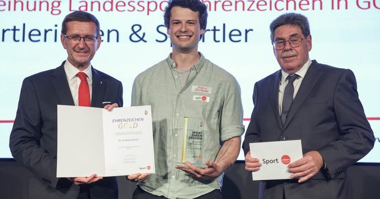 Drei Männer stehen zusammen, einer hält ein Papier, ein anderer einen Pokal, während ein Dritter lächelt. Der Hintergrund zeigt eine große Leinwand mit dem Text 'Landessportehrenzeichen & Sportler'.