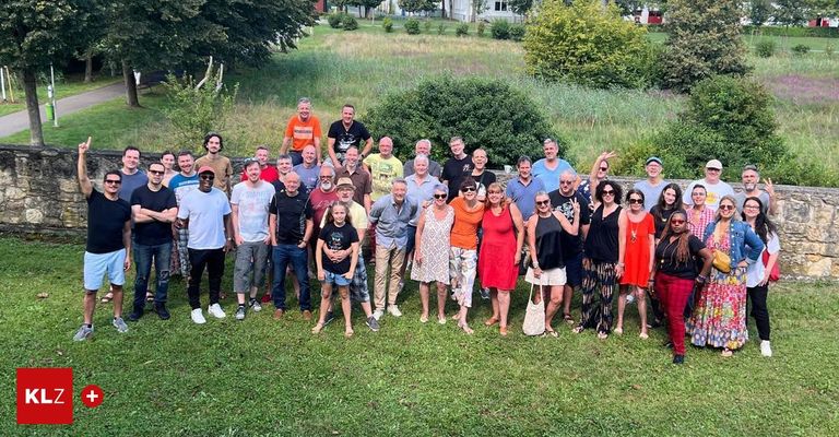 Eine Gruppe von Menschen steht gemeinsam auf einem Rasenfeld, lächelt und posiert für ein Foto. Sie sind lässig gekleidet, einige tragen Sonnenbrillen. Hinter ihnen befindet sich eine üppige Landschaft mit verschiedenen Pflanzen und Bäumen.