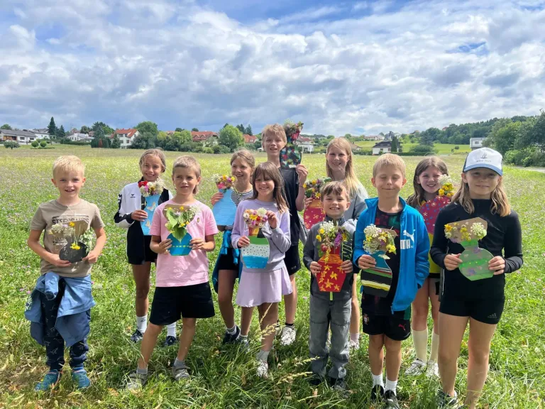 Eine Gruppe von Kindern steht in einem Feld und hält selbstgemachte Blumensträuße. Sie lächeln für die Kamera. Der Himmel ist bewölkt.