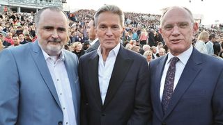 Drei Männer in Anzügen stehen vor einer großen Menge, möglicherweise bei einer Veranstaltung.