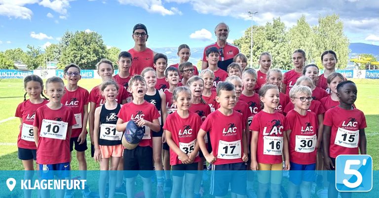 Eine Gruppe von Kindern in roten Shirts mit Nummern, wahrscheinlich bei einem Rennen, posiert für ein Foto. Zwei Männer stehen hinter ihnen, einer trägt eine Brille und eine Uhr.