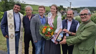 Eine Gruppe von Menschen steht im Freien. Eine Frau mit Krone hält einen Blumenstrauß. Ein Mann hält eine Flasche. Hinter ihnen steht ein Haus und ein Feld.