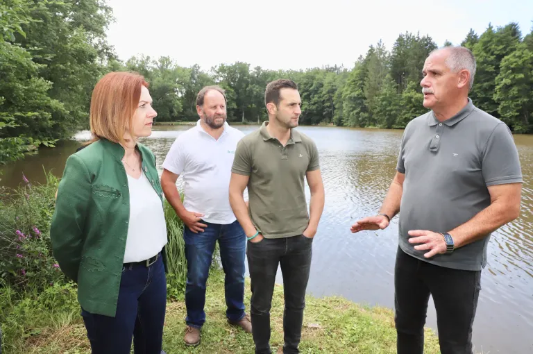 Vier Erwachsene stehen in der Nähe eines Sees mit Bäumen im Hintergrund. Die Frau links trägt eine grüne Jacke, während die anderen Poloshirts tragen. Sie scheinen über etwas zu diskutieren.