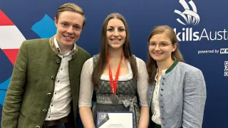 Drei Menschen stehen mit einem Zertifikat vor einer blauen Wand. Eine Frau in der Mitte trägt eine Medaille und hält ein Zertifikat. Die Frau auf der rechten Seite trägt eine Brille. Hinter ihnen ist ein Logo mit dem Text 'skills'.