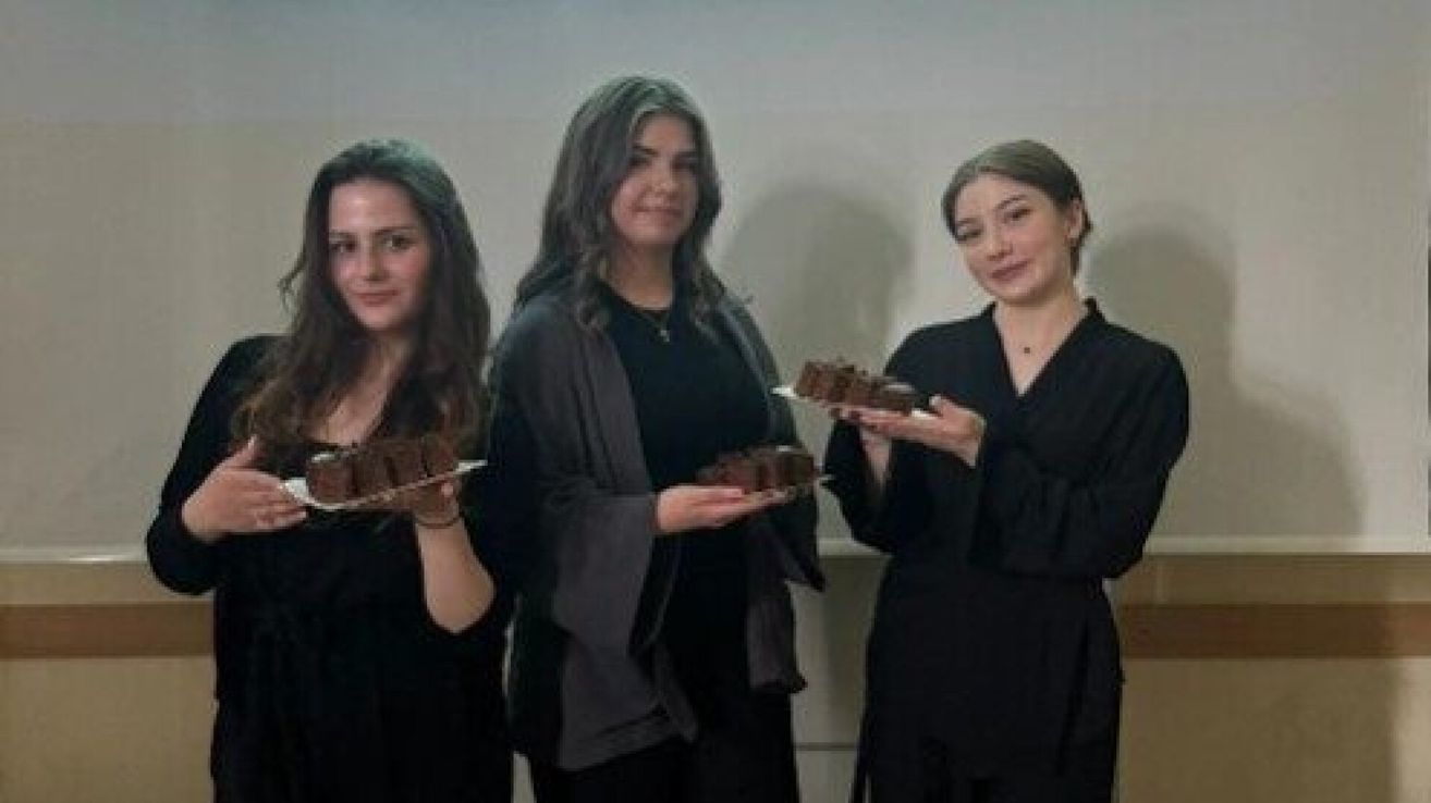 Drei Frauen in schwarzen Kleidern stehen in einer Reihe und halten Teller mit Schokoladendesserts. Die Frau auf der linken Seite lächelt, während die anderen für ein Foto posieren.