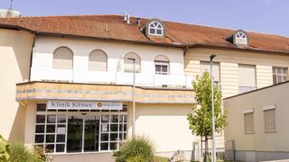 Ein beigefarbenes Gebäude mit einem braunen Dach hat mehrere Fenster, einen Balkon und ein Schild mit der Aufschrift 'ik Kittsee burgenland'. Vor dem Gebäude stehen zwei Straßenlaternen und ein Baum.