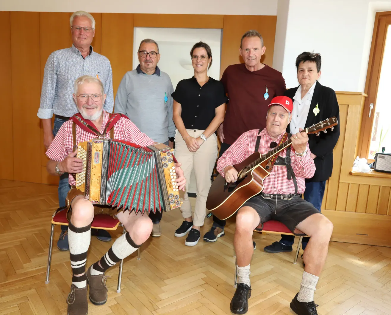Eine Gruppe von Menschen steht und sitzt in einem Raum. Zwei Männer spielen Musikinstrumente, einer spielt Akkordeon und der andere Gitarre. Hinter ihnen stehen drei weitere Personen und lächeln. Der Raum hat Holzböden und Holzwände.
