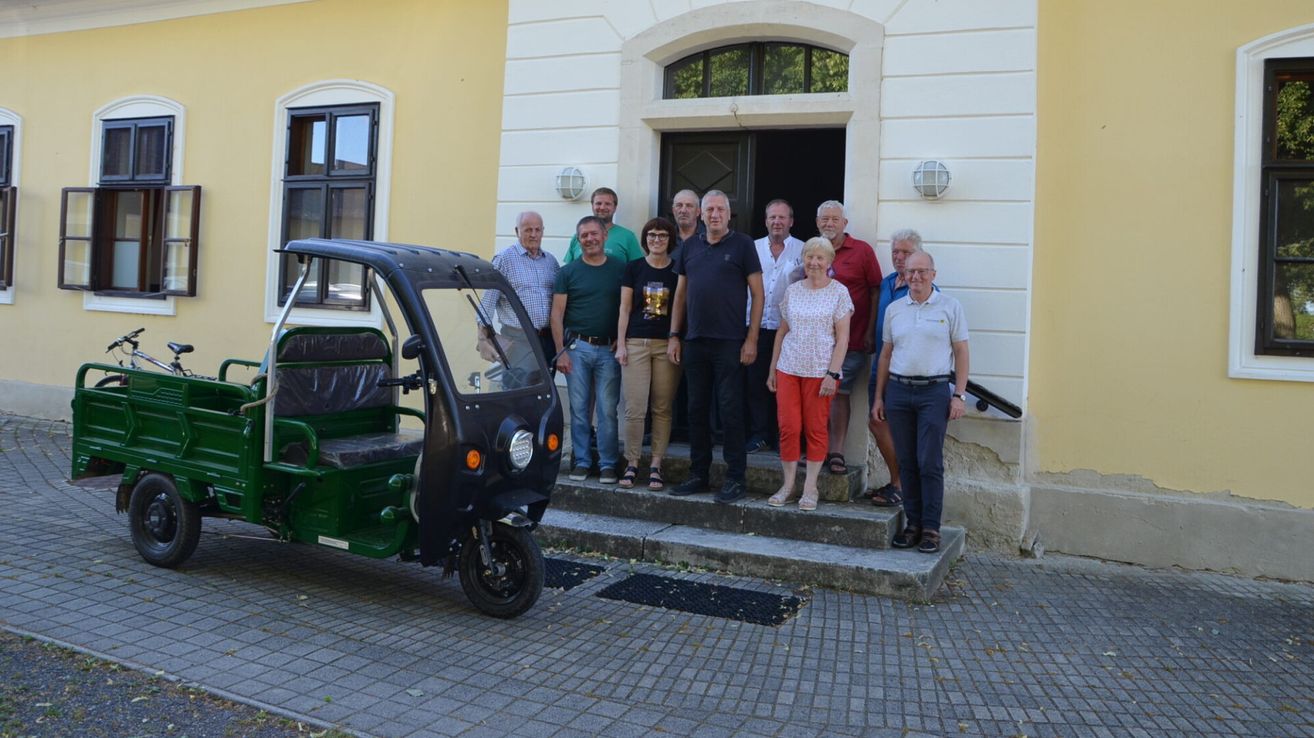Eine Gruppe von Menschen steht auf den Stufen eines Gebäudes, mit einem kleinen grünen Fahrzeug davor geparkt. Sie lächeln und scheinen für ein Foto zu posieren.