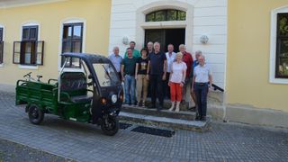 Eine Gruppe von Menschen steht auf den Stufen eines Gebäudes, mit einem kleinen grünen Fahrzeug davor geparkt. Sie lächeln und scheinen für ein Foto zu posieren.