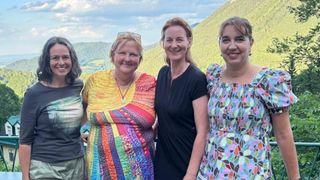 Vier Frauen stehen zusammen und lächeln fröhlich, vor einer bergigen Kulisse. Eine trägt ein leuchtend mehrfarbiges Kleid, während die anderen blumige und dunkelfarbige Kleider tragen.