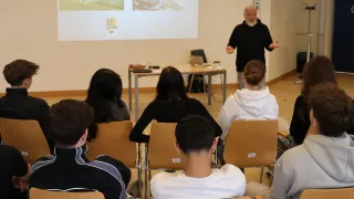 Ein Klassenzimmer mit einem Mann, der eine Präsentation hält. Eine Projektionsleinwand zeigt ein Bild, und das Publikum sitzt aufmerksam. Der Mann steht mit offenen Armen und gestikuliert in Richtung der Leinwand.
