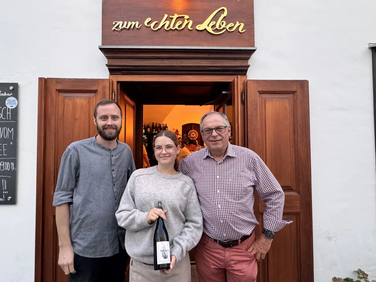 Drei Personen stehen vor einer Holztür mit dem Schriftzug 'zum echten Leben'. Eine Frau hält eine Flasche, während ein Mann links von ihr ein graues Hemd trägt und der Mann rechts von ihr ein kariertes Hemd und eine Brille trägt.