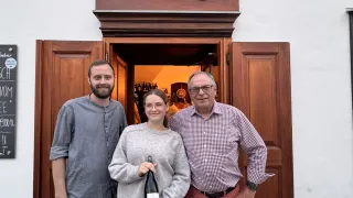 Drei Personen stehen vor einer Holztür mit dem Schriftzug 'zum echten Leben'. Eine Frau hält eine Flasche, während ein Mann links von ihr ein graues Hemd trägt und der Mann rechts von ihr ein kariertes Hemd und eine Brille trägt.