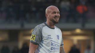 Ein Mann in einem weißen und grauen Fußballtrikot lächelt und posiert für die Kamera. Das Trikot hat blauen Text und ein gelbes Abzeichen auf der Schulter. Der Hintergrund ist unscharf.