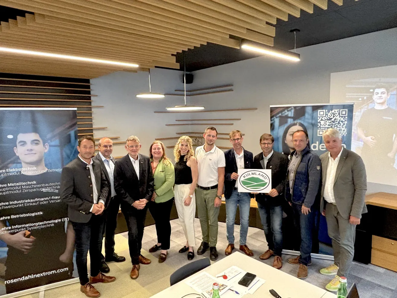 Eine Gruppe von Fachleuten posiert für ein Foto in einem Büro. Sie stehen in einer Reihe, wobei ein Mann ein Schild hält. Ein Tisch und Stühle stehen vor ihnen. Eine Wand mit Postern ist hinter ihnen.