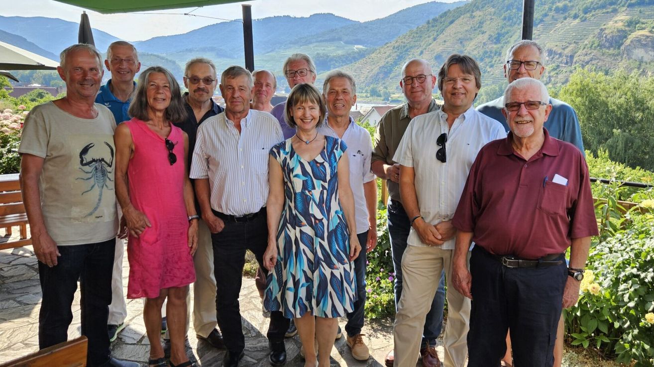 Eine Gruppe Erwachsener lächelt und posiert für ein Foto, umgeben von einer Berglandschaft. Sie sind in Freizeitkleidung gekleidet, einige tragen Brillen und Accessoires.