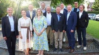 Eine Gruppe von Menschen, darunter Männer und Frauen, stehen lächelnd und posieren für ein Foto auf einem gepflasterten Bürgersteig. Sie tragen formelle Kleidung.