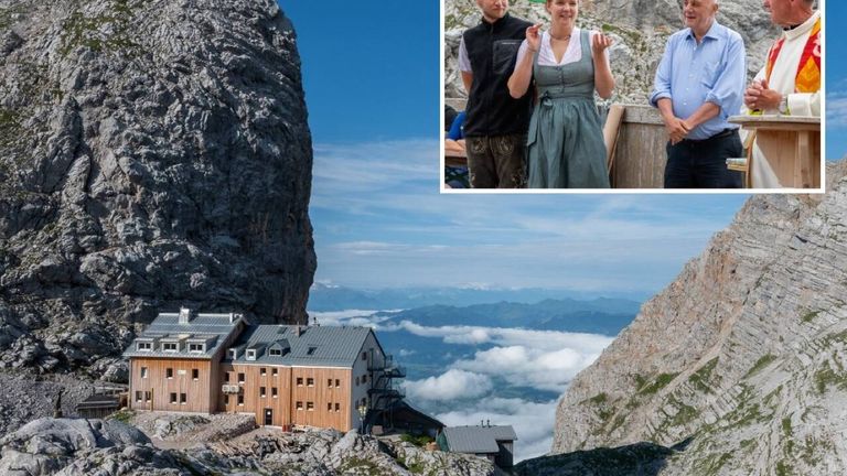 Ein Mann und eine Frau in traditioneller bayerischer Kleidung posieren für ein Foto vor einem Berg. Unten steht ein Haus auf einem felsigen Felsen mit Bergen im Hintergrund.