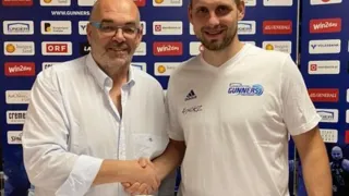 Zwei Männer stehen vor einem Tisch mit einem Basketball. Sie lächeln und schütteln Hände. Hinter ihnen ist eine Wand mit verschiedenen Logos und Sponsoren.