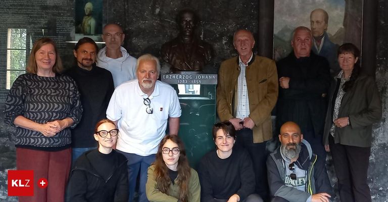 Eine Gruppe von Menschen posiert vor einer Büste von Erzherzog Johann. Sie lächeln und stehen um eine grüne Plakette herum.
