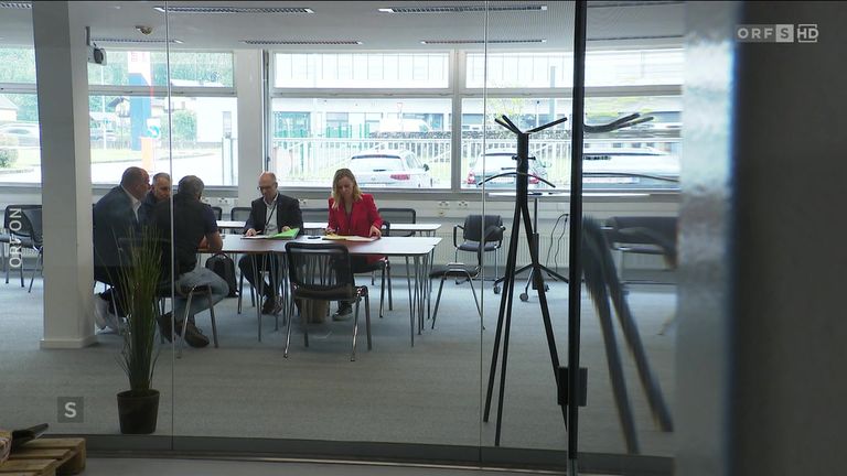 Ein Büroraum mit Glaswänden. Eine Frau und ein Mann sitzen an einem Tisch. Sie scheinen eine Diskussion zu führen. Die Glaswand spiegelt einen anderen Raum mit Stühlen und einem Kleiderständer wider.