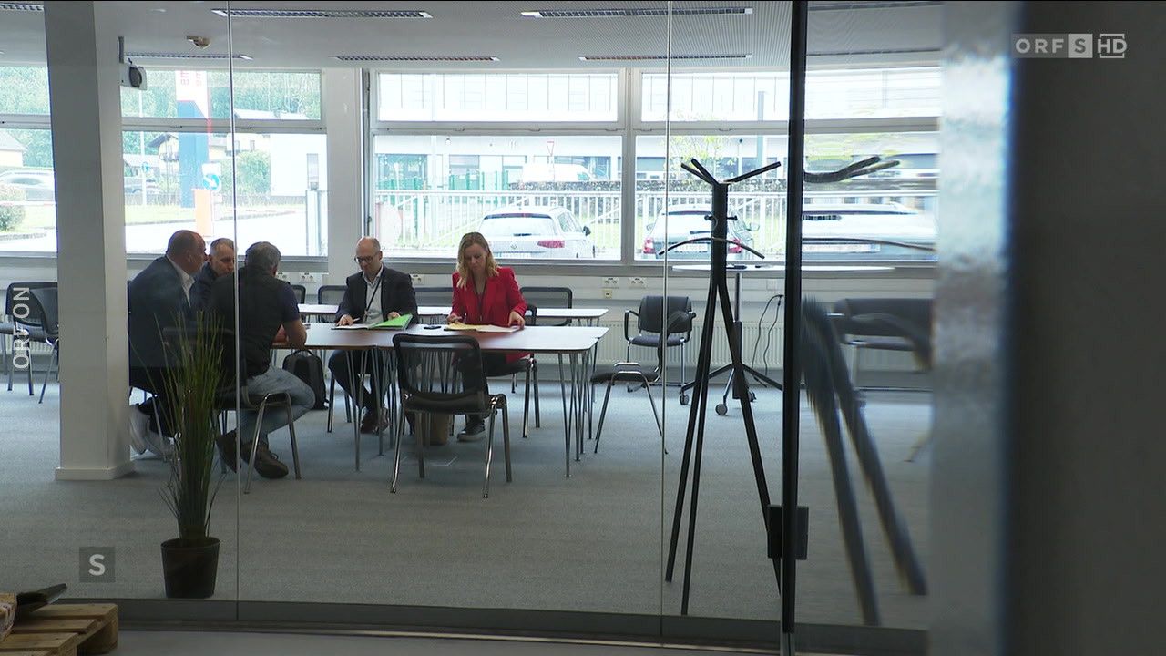 Ein Büroraum mit Glaswänden. Eine Frau und ein Mann sitzen an einem Tisch. Sie scheinen eine Diskussion zu führen. Die Glaswand spiegelt einen anderen Raum mit Stühlen und einem Kleiderständer wider.