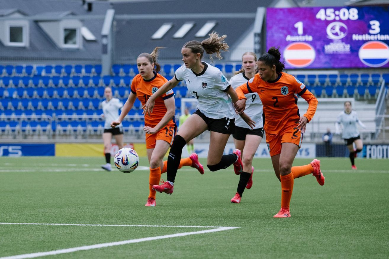 Frauenfußballspielerinnen in orangefarbenen und weißen Trikots spielen auf einem Feld. Die Spielerin in Weiß tritt den Ball. Die Anzeigetafel im Hintergrund zeigt Österreich mit 1:0 in Führung.