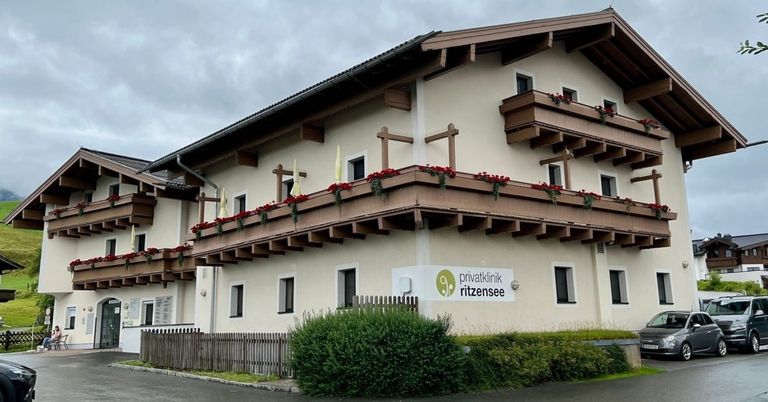 Die Vorderseite eines Gebäudes mit roten Blumen auf Balkonen, einem Schild mit der Aufschrift 'privatklinik ritzensee' und einem Zaun davor.