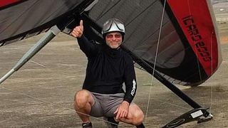 Ein Mann in einer schwarzen Jacke und grauen Shorts hockt neben einem roten und schwarzen Paraglider und gibt einen Daumen hoch.