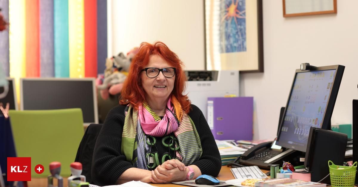 Eine Frau mit roten Haaren, die eine Brille und einen Schal trägt, sitzt an einem Schreibtisch mit einem Computer, Büchern und einem Telefon. Hinter ihr befindet sich eine Regenbogenwand und ein gerahmtes Bild.