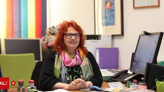 Eine Frau mit roten Haaren, die eine Brille und einen Schal trägt, sitzt an einem Schreibtisch mit einem Computer, Büchern und einem Telefon. Hinter ihr befindet sich eine Regenbogenwand und ein gerahmtes Bild.