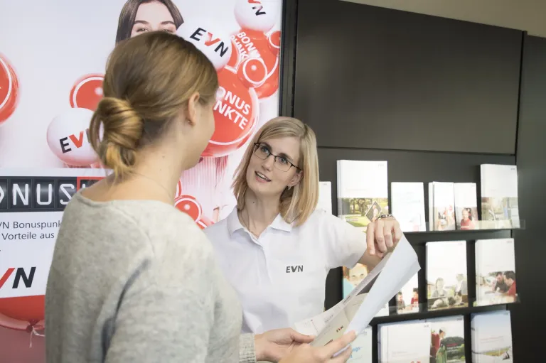 Eine Frau mit Brille hält ein Papier und spricht mit einer anderen Frau in einem Geschäft. Dahinter befindet sich ein Schild mit dem Wort EVN und Bällen mit roten Buchstaben.