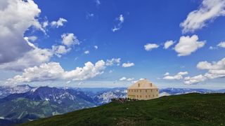 Ein Haus auf einem Berggipfel mit einer Gruppe von Menschen davor, umgeben von einem grasigen Feld und majestätischen Bergen unter einem blauen Himmel mit Wolken.