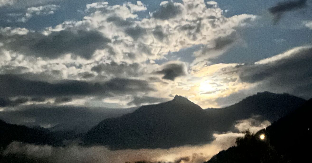 Ein Gebirgszug mit Nebel darunter unter einem teilweise bewölkten Himmel, durch den die Sonne lugt.