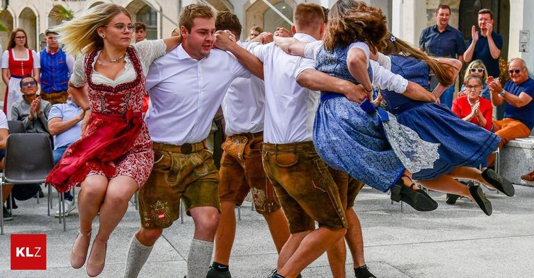 Eine Gruppe von Menschen in traditioneller deutscher Kleidung tanzt im Freien im Kreis. Eine Frau wird von zwei Männern hochgehoben, während andere mittanzen.