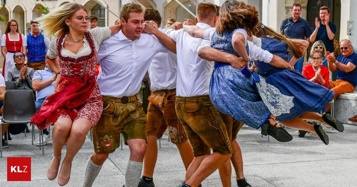 Eine Gruppe von Menschen in traditioneller deutscher Kleidung tanzt im Freien im Kreis. Eine Frau wird von zwei Männern hochgehoben, während andere mittanzen.