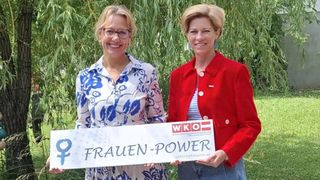 Zwei Frauen stehen vor einem Hintergrund von Grünpflanzen und lächeln. Die Frau auf der linken Seite trägt eine Brille und eine gemusterte Bluse. Die Frau auf der rechten Seite trägt einen roten Blazer und hält ein weißes Banner mit der Aufschrift 'FRAUEN-POWER'.