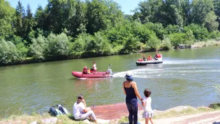 Zwei Boote auf einem Fluss mit Menschen darin. Eine Frau und ein Kind beobachten vom Ufer aus.