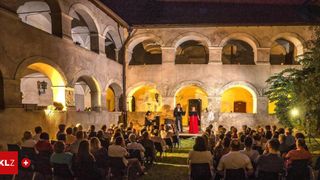 Eine nächtliche Aufführung unter einem alten Innenhof mit Bogenfenstern. Ein Mann und eine Frau singen auf der Bühne, während ein Publikum in Stühlen um sie herum sitzt.