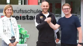 Zwei Männer schütteln sich vor einem Gebäude mit einem Schild, auf dem 'Berufsschule' steht. Sie lächeln. Ein grünes Schild befindet sich hinter ihnen.