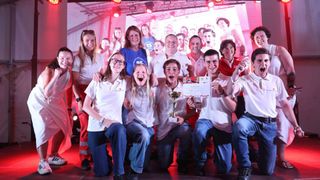 Eine Gruppe lächelnder Personen in weißen und roten Uniformen, wahrscheinlich Freiwillige, posiert für ein Foto mit Trophäen und Zertifikaten auf einer rot beleuchteten Bühne.
