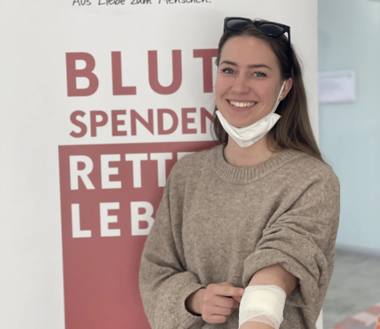 Eine Frau mit Gesichtsmaske und Pullover steht lächelnd vor einem Schild, das 'BLUT SPENDEN RETTE LEB' besagt. Sie hat einen Verband am Arm.