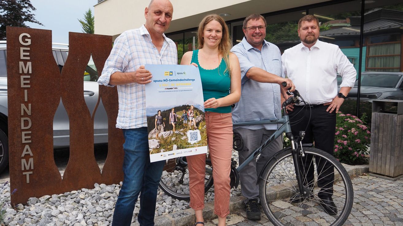 Vier Personen stehen vor einem Gebäude, einer hält ein Plakat, der andere ein Fahrrad. Das Plakat zeigt eine Gruppe von Wanderern und einen QR-Code.