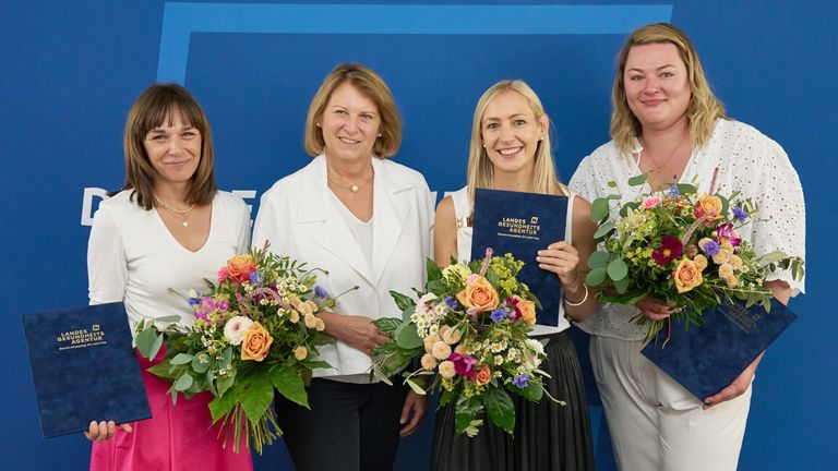 Vier Frauen stehen lächelnd nebeneinander und halten Blumen. Die Frau in der Mitte hält ein blaues Zertifikat. Sie stehen vor einem blauen Hintergrund.