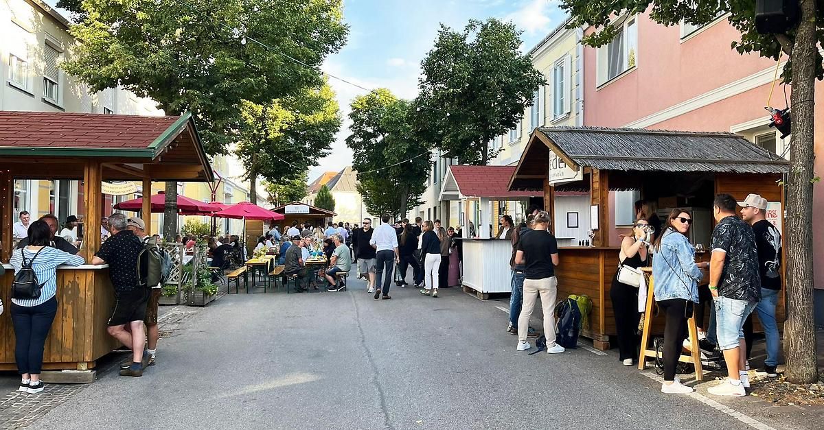 Ein geschäftiger Straßenmarkt mit Menschen, die unter Schirmen an Tischen sitzen. Einige Leute gehen vorbei, und einige stehen an einem kleinen Holztresen. Bäume säumen die Straße.