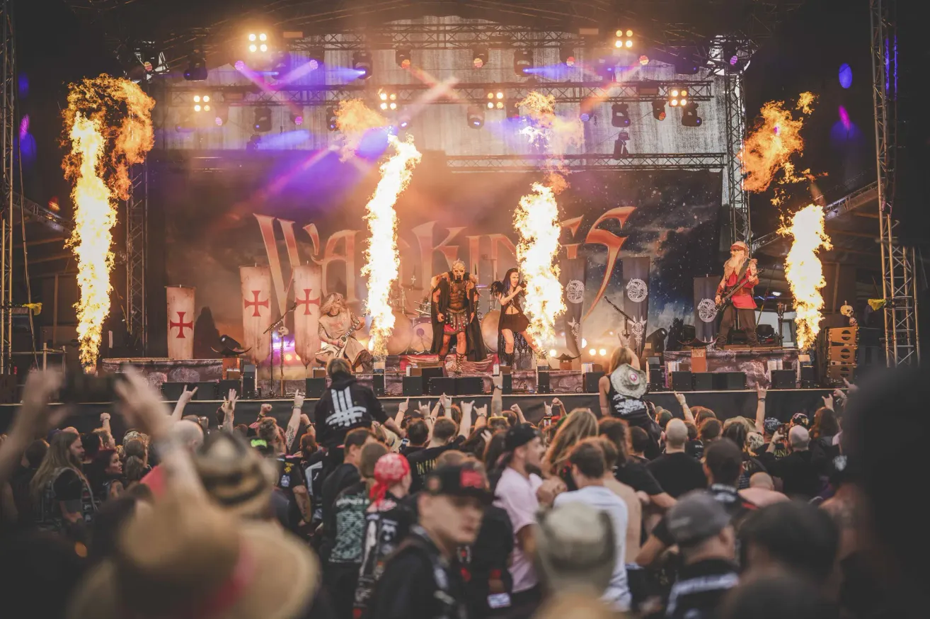 Eine Heavy-Metal-Band tritt mit Flammen, unter Scheinwerfern und umgeben von einer Menge auf der Bühne auf.